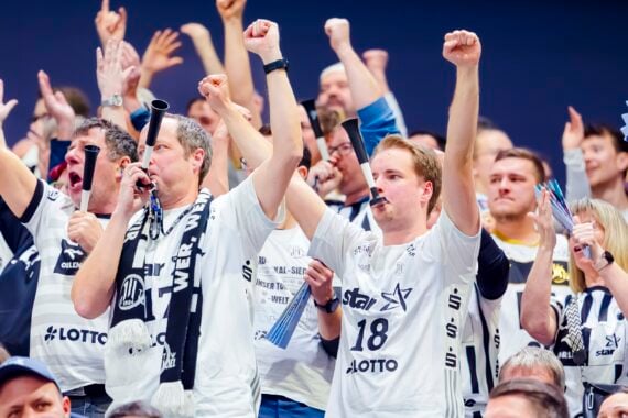 Eine Menge begeisterter Sportfans in einheitlichen weißen Trikots jubelt und reckt bei einer Hallenveranstaltung im Gleichklang die Fäuste in die Höhe – ein Bild, das die elektrisierende Atmosphäre des ausverkauften Derbys zwischen Kiel und Flensburg in der Handball-Bundesliga einfängt.
