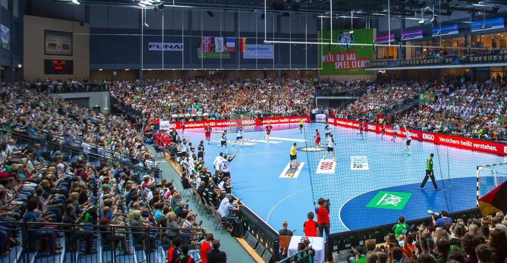 Eine große Sporthalle voller Zuschauer, die ein Handballspiel auf einem blauen Spielfeld verfolgen, während die Spieler in Bewegung sind und über ihnen bunte Banner hängen.