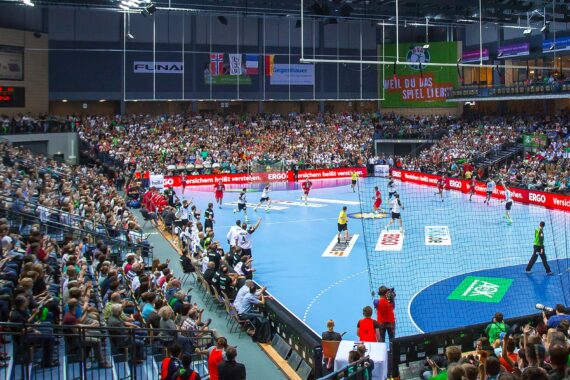 Eine große Sporthalle voller Zuschauer, die ein Handballspiel auf einem blauen Spielfeld verfolgen, während die Spieler in Bewegung sind und über ihnen bunte Banner hängen.