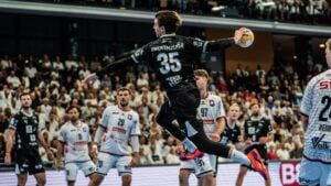 Ein Handballspieler in schwarzer Kleidung springt mit dem Ball in der Hand in die Luft, um ein Tor zu erzielen, während Spieler in weißer Kleidung verteidigen. Die Halle ist voller Zuschauer, die gebannt zuschauen.