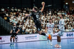 Ein Handballspieler in schwarzer Spielkleidung springt hoch, um einen Wurf auszuführen, während Spieler in weißer Spielkleidung zuschauen. Im Hintergrund ist eine Zuschauermenge in einer hell beleuchteten Sporthalle zu sehen.