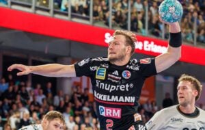 Ein Handballspieler in einem schwarzen Trikot springt während eines Spiels hoch, um den Ball zu werfen.