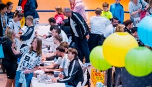Sportler einer Mannschaft geben an Tischen in einer Sporthalle Autogramme für Fans.