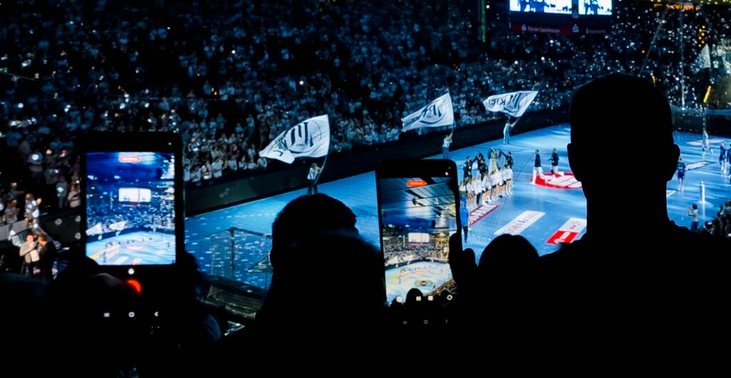 Zuschauer in einer abgedunkelten Arena halten ihre Smartphones hoch, um ein Handballspiel auf dem blauen Spielfeld unter ihnen aufzunehmen.