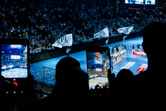 Zuschauer in einer abgedunkelten Arena halten ihre Smartphones hoch, um ein Handballspiel auf dem blauen Spielfeld unter ihnen aufzunehmen.