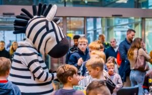 Eine Person in einem Zebra-Maskottchenkostüm unterhält sich mit einer Gruppe von Kindern in einer Lobby.
