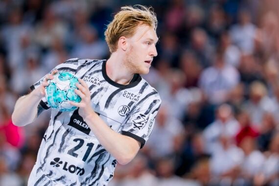 Ein Handballspieler in einem schwarz-weißen Trikot hält einen blau-weißen Ball in der Hand und blickt nach links. Im Hintergrund ist eine verschwommene Menschenmenge zu sehen, was auf eine Hallensportveranstaltung hindeutet.