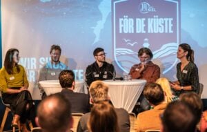 Eine Diskussionsrunde aus vier Personen, die an zwei runden Tischen auf einer Bühne vor einer Leinwand mit dem THW-Kiel-Logo sitzen.
