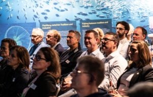 Das Publikum sitzt in Reihen und verfolgt einen Vortrag vor einem Hintergrund, auf dem ein Fischschwarm und der Schriftzug Fischen zwischen Tradition und Industrie zu sehen sind.