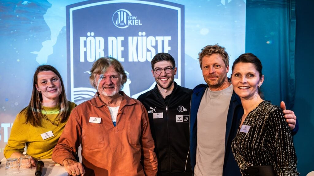 26_Foer_De_Kueste_B2B_Titel Fünf Personen posieren für ein Foto vor einem blauen Veranstaltungshintergrund des THW Kiel För de Küste.