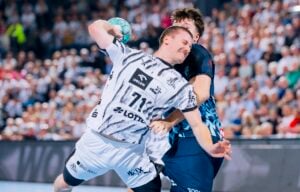 Ein Handballspieler im weißen Trikot macht sich bereit, den Ball zu werfen, während er von einem gegnerischen Spieler im dunkelblauen Trikot geblockt wird.