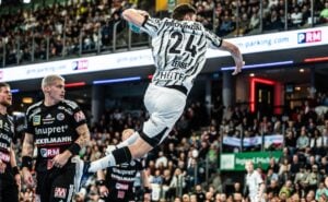 Ein Handballspieler in einem weißen Trikot springt während eines Spiels in einer vollbesetzten Halle in die Luft, um den Ball zu werfen.