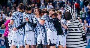 Eine Handballmannschaft in weiß-schwarz gemusterten Trikots steht eng beieinander neben einem Zebra-Maskottchen.