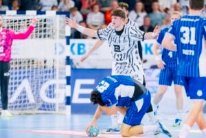 Ein Handballspieler in blauer Kleidung beugt sich mit dem Ball in der Hand vor, während hinter ihm ein Spieler in einem schwarz-weiß gestreiften Trikot mit erhobenen Armen steht. Im Hintergrund sind das Tor und weitere Spieler zu sehen.