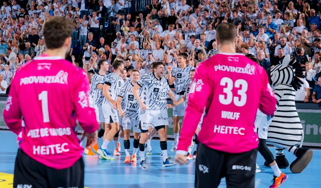 HANDBALL 2026/27: THW Kiel - SG Flensburg-Handewitt Eine Handballmannschaft in weißen Trikots feiert auf dem Spielfeld zusammen mit zwei Torhütern in rosa Trikots und einem Zebra-Maskottchen, während die Fans im Hintergrund jubeln.