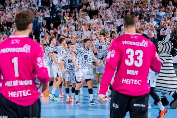 Eine Handballmannschaft in weißen Trikots feiert auf dem Spielfeld zusammen mit zwei Torhütern in rosa Trikots und einem Zebra-Maskottchen, während die Fans im Hintergrund jubeln.