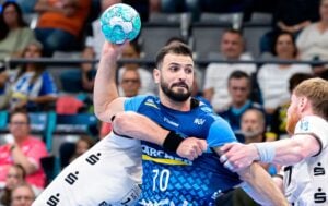 Ein Handballspieler im blauen Trikot macht sich bereit, einen blau gemusterten Ball zu werfen, während er von zwei Gegnern in weißen Trikots geblockt wird.