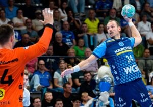 Ein Handballspieler in einem blauen Trikot springt hoch, um den Ball in Richtung eines Torwarts in einem orangefarbenen Trikot zu werfen.