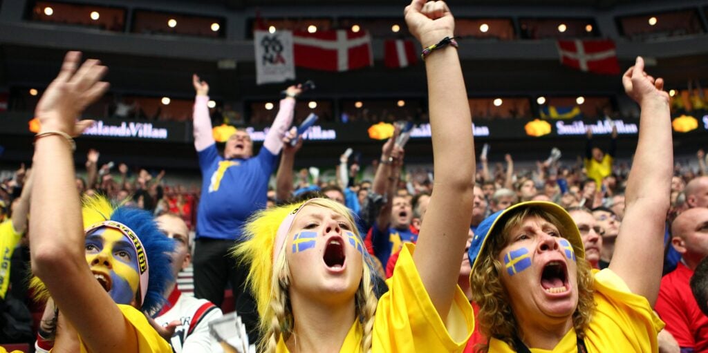 Schwedische Sportfans in gelben Trikots und mit Gesichtsbemalung, die auf den Tribünen mit erhobenen Armen jubeln.