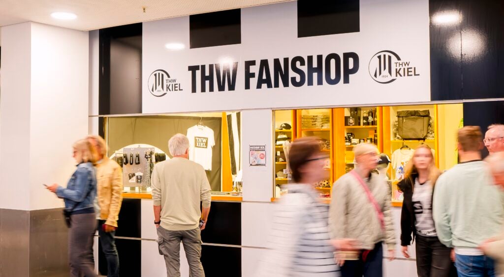 Einkäufer gehen am Fanshop des THW Kiel vorbei, in dessen Schaufenster Mannschaftskleidung und Fanartikel ausgestellt sind.