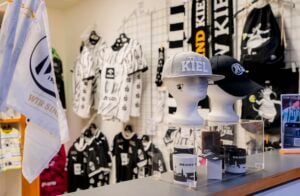 Zwei Schaufensterpuppenköpfe mit THW-Kiel-Baseballkappen sitzen auf einer Glasvitrine vor Markenschals, Trikots und Tassen.