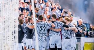 Fußballspieler in gemusterten weißen Trikots feiern ein Tor, während jubelnde Fans von den Tribünen aus zuschauen.