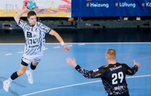 Ein Handballspieler in einem weißen Trikot springt hoch, um den Ball in Richtung eines Torhüters in einem schwarzen Trikot mit der Nummer 29 zu werfen.