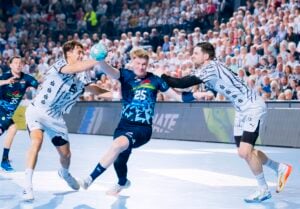 Ein Handballspieler im blauen Trikot springt hoch, um den Ball zu werfen, während er von zwei Gegnern in weißen Trikots verteidigt wird.