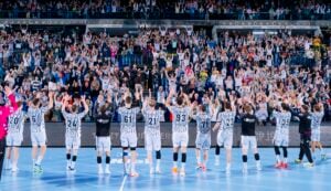 Eine Handballmannschaft in weiß-schwarzen Trikots steht mit dem Rücken zur Kamera und hat die Arme erhoben, um einer großen, jubelnden Zuschauermenge auf den Tribünen zu applaudieren.