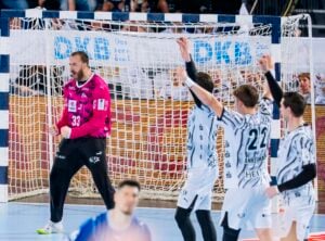 Ein Handballtorwart in einem rosa Trikot steht aufmerksam neben dem Tor, während die Spieler der gegnerischen Mannschaft in weißen und schwarzen Trikots jubelnd die Arme in die Höhe recken. Im Hintergrund sind das Tor und ein Teil der Sporthalle zu sehen.