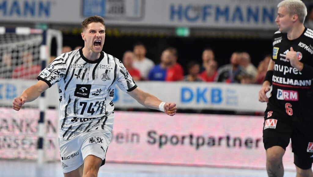 Ein Handballspieler in einem schwarz-weißen Trikot jubelt jubelnd auf dem Spielfeld, während ein anderer Spieler in einem schwarzen Trikot daneben steht und zusieht.