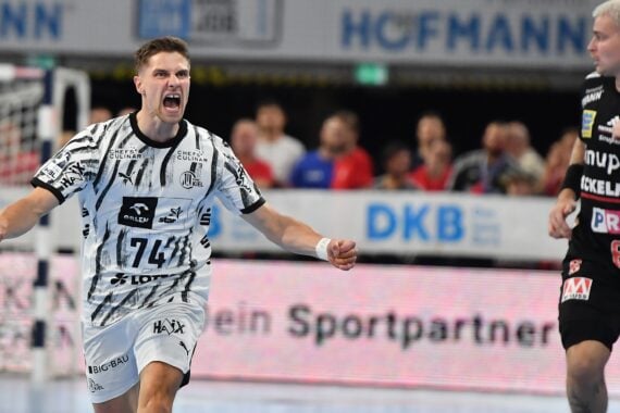Ein Handballspieler in einem schwarz-weißen Trikot jubelt jubelnd auf dem Spielfeld, während ein anderer Spieler in einem schwarzen Trikot daneben steht und zusieht.