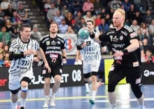 Ein Handballspieler in einem schwarzen Trikot macht sich während eines Spiels bereit, den Ball zu werfen, während seine Mannschaftskameraden zuschauen.