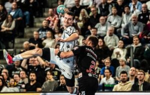 Ein Handballspieler im weißen Trikot springt hoch, um den Ball zu werfen, während er von einem Verteidiger im schwarzen Trikot während eines Spiels vor einer vollbesetzten Halle geblockt wird.