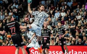 Ein Handballspieler in einem weißen Trikot springt hoch, um den Ball zu werfen, umgeben von Mitspielern und Gegnern in einer überfüllten Halle.