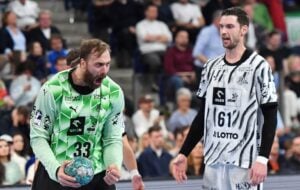 Zwei Handballspieler in Mannschaftstrikots, einer in Grün und einer in Schwarz-Weiß, stehen auf einem Spielfeld vor einer Zuschauermenge.