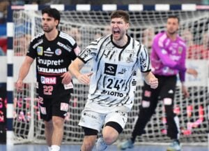Ein Handballspieler in einem schwarz-weißen Trikot jubelt auf dem Spielfeld, im Hintergrund sind ein gegnerischer Spieler und ein Torwart zu sehen.