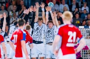 Zwei Handballspieler in weißen Trikots springen hoch, um einen Wurf vor dem Tor abzuwehren, während Spieler in roten Trikots zusehen.