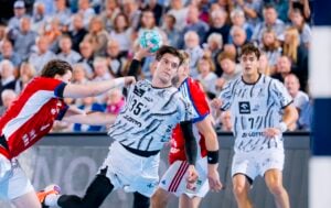 Ein Handballspieler in einem weiß gemusterten Trikot springt hoch, um den Ball zu werfen, während er von Spielern in roten und weißen Trikots verteidigt wird.