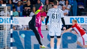 Ein Handballtorwart springt hoch, um einen Ball in der Nähe des Tors abzuwehren, während zwei andere Spieler zusehen.
