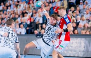 Ein Handballspieler in einem schwarz-weiß gemusterten Trikot springt hoch, um den Ball an einem Verteidiger in einem rot-weiß-blauen Trikot vorbei zu werfen.