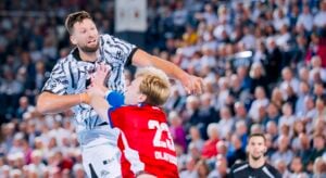 Ein Handballspieler in einem weiß gemusterten Trikot wird von einem Gegner in einem roten Trikot mit der Rückennummer 23 geblockt.