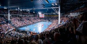 Eine Aufnahme aus großer Höhe einer vollbesetzten Handballhalle, in der die Spieler auf einem blauen Spielfeld gegeneinander antreten.