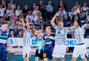 Vier männliche Handballspieler in weißen und violetten Trikots feiern auf dem Spielfeld vor den sitzenden Zuschauern.