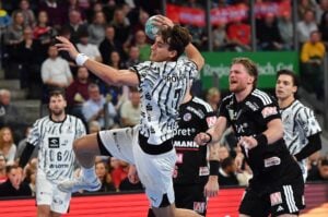 Ein Handballspieler in einem weiß gemusterten Trikot springt in der Luft hoch, um den Ball zu werfen, während ein Verteidiger in Schwarz versucht, ihn daran zu hindern.