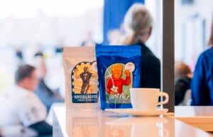 Zwei Kaffeebeutel, einer in Weiß und einer in Blau, mit Abbildungen von Handballspielern, stehen auf einer Theke neben einer weißen Kaffeetasse.