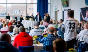 Ein Raum voller Menschen, die einer Präsentation beiwohnen; im Vordergrund steht ein Redner am Podium und ein Mann, der ein Handballtrikot mit den Aufschriften PROVINZIAL und WOLFF auf dem Rücken trägt.