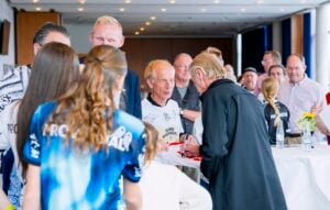 Horst Hrubesch, der eine schwarze Jacke trägt, signiert bei einer Veranstaltung ein weißes Sporttrikot für eine Gruppe von Fans.