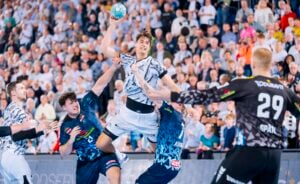Ein Handballspieler springt in die Luft, um den Ball zu werfen, während er vor den Augen der Stadionzuschauer von zwei gegnerischen Spielern geblockt wird.