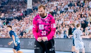 Ein Handballtorwart in einem knallrosa Trikot jubelt mit geballten Fäusten während eines Spiels in einer vollbesetzten Halle.